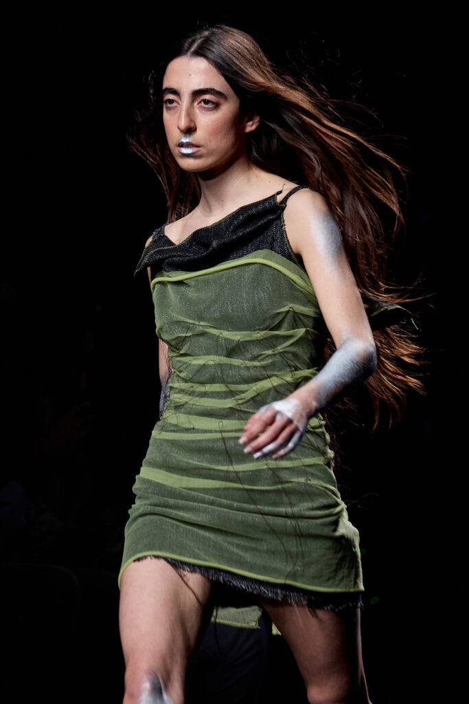 Una modelo camina por la pasarela luciendo un vestido verde con detalles oscuros y un maquillaje distintivo.