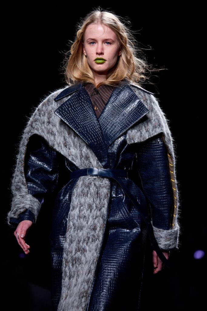 Modelo desfilando con un abrigo de cuero y pelo, con labios pintados de verde.