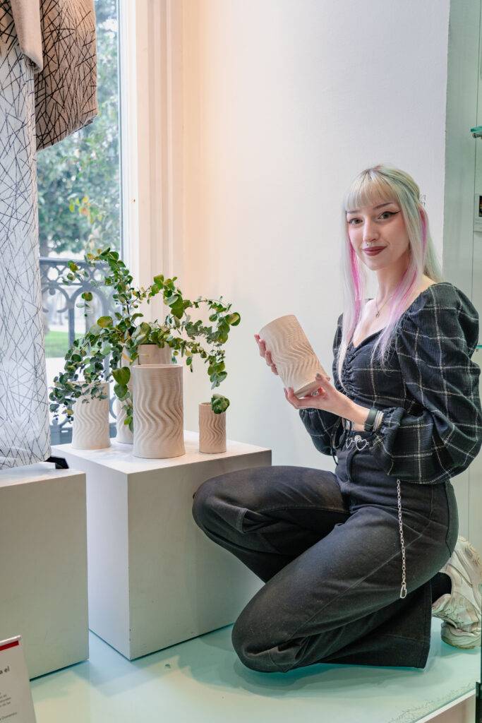 Una mujer con cabello de tonos pastel sostiene un jarrón en un entorno decorativo con plantas.