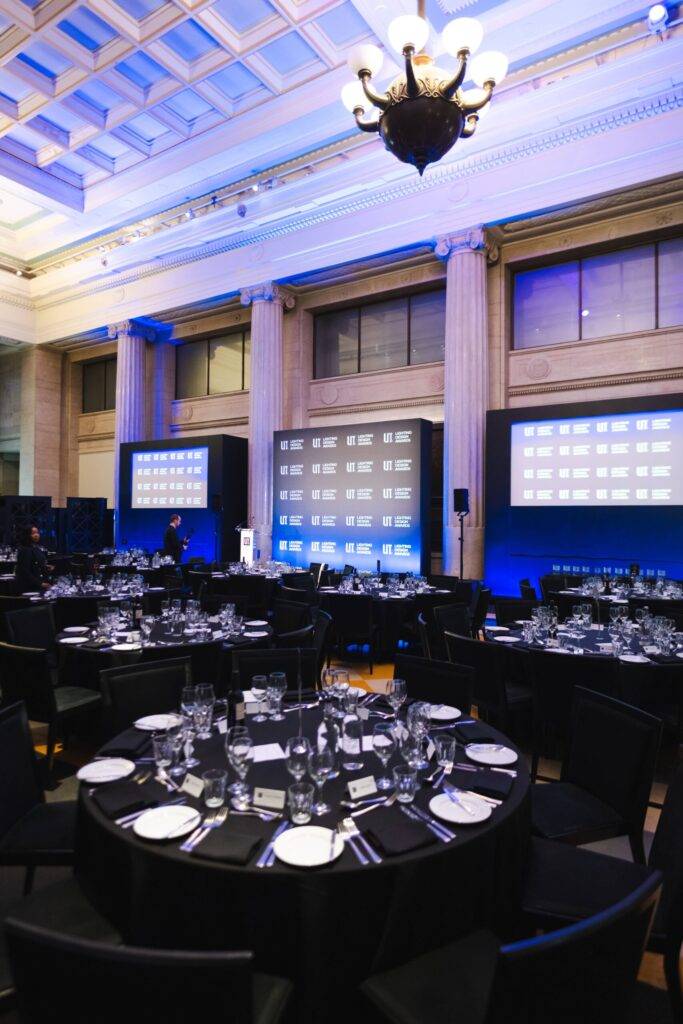 La imagen muestra un elegante salón con mesas preparadas para un evento formal, iluminado con tonos azules.