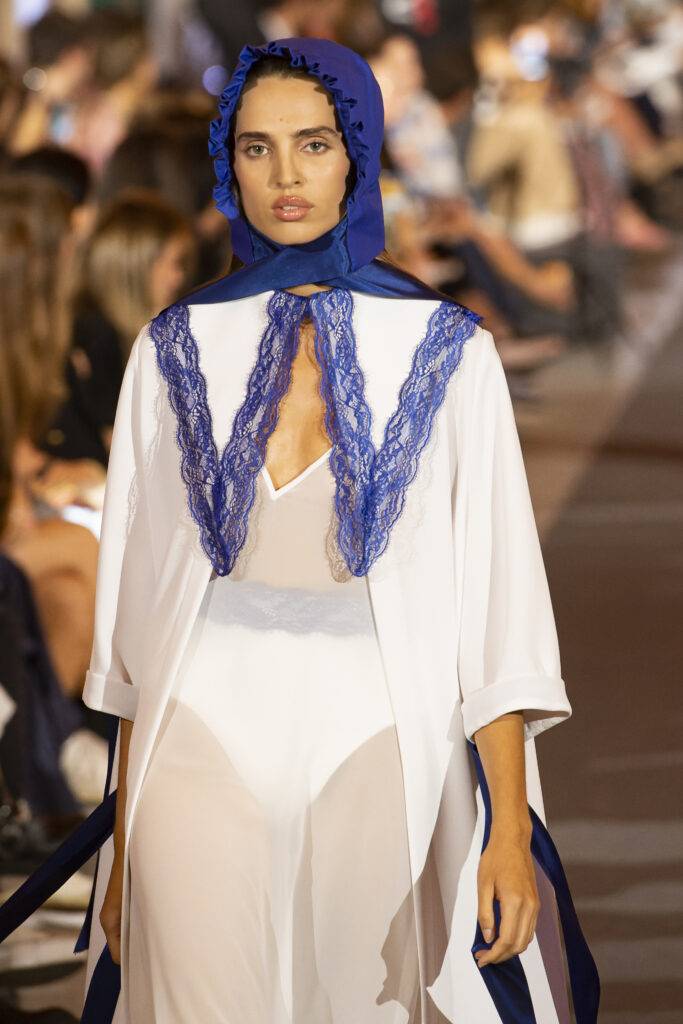 Modelo desfilando con un atuendo blanco decorado con encaje azul en un evento de moda.