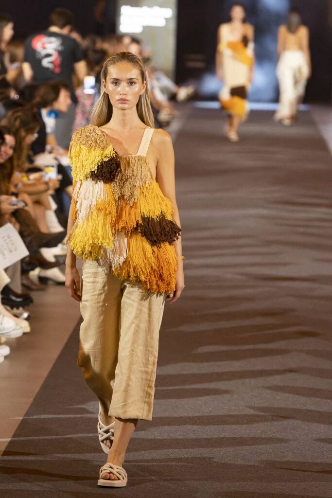 Una modelo camina por la pasarela luciendo un conjunto de moda con flecos en tonos amarillos y marrones.