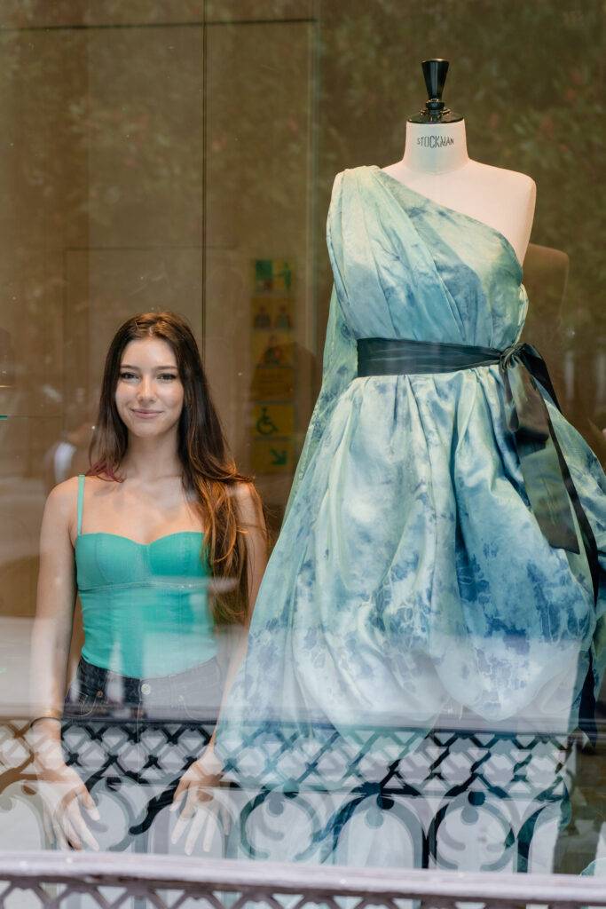 Una joven sonriente se encuentra frente a un vestido largo expuesto en una vitrina.