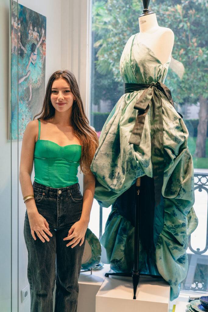 Una joven posa junto a un maniquí que exhibe un vestido verde con un diseño elegante.