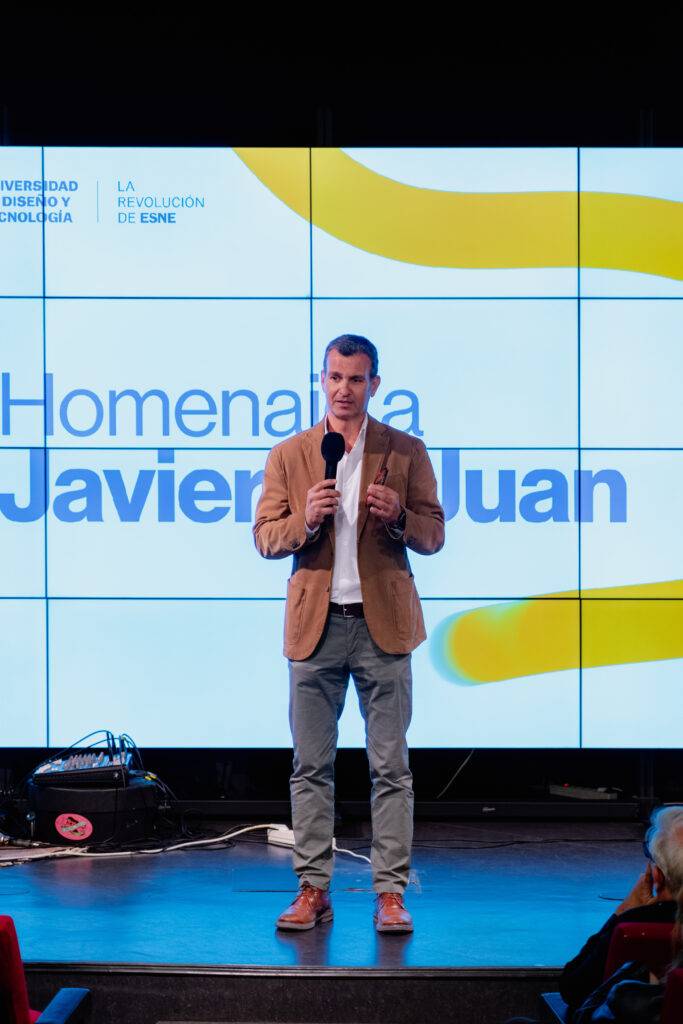 Una persona está hablando en un escenario frente a una pantalla con un mensaje de homenaje.