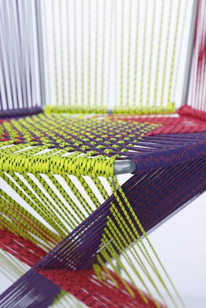 La imagen muestra un diseño moderno y colorido de una silla hecha con cuerdas metálicas y plásticas en tonos vibrantes.