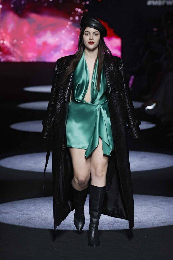 Una modelo camina por la pasarela luciendo un atuendo elegante en tonos verdes y negros.