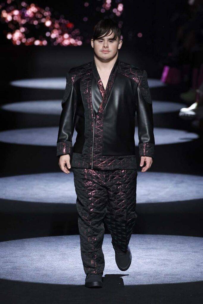Un modelo camina por la pasarela luciendo un traje de cuero negro con detalles en color púrpura.