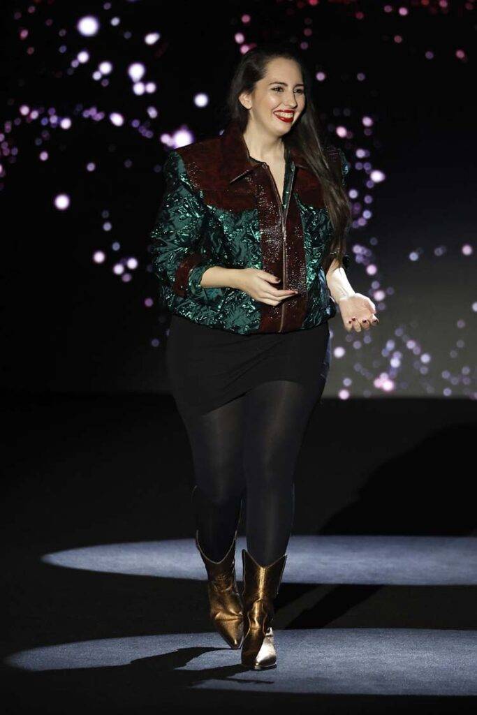 Una mujer camina con confianza en un evento, luciendo un atuendo llamativo con una chaqueta brillante y botas doradas.