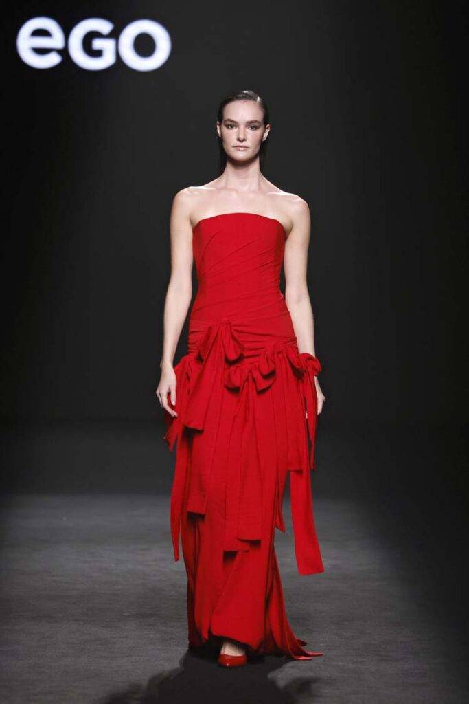 Una modelo camina con un vestido largo y rojo adornado con pliegues y cintas en una pasarela.