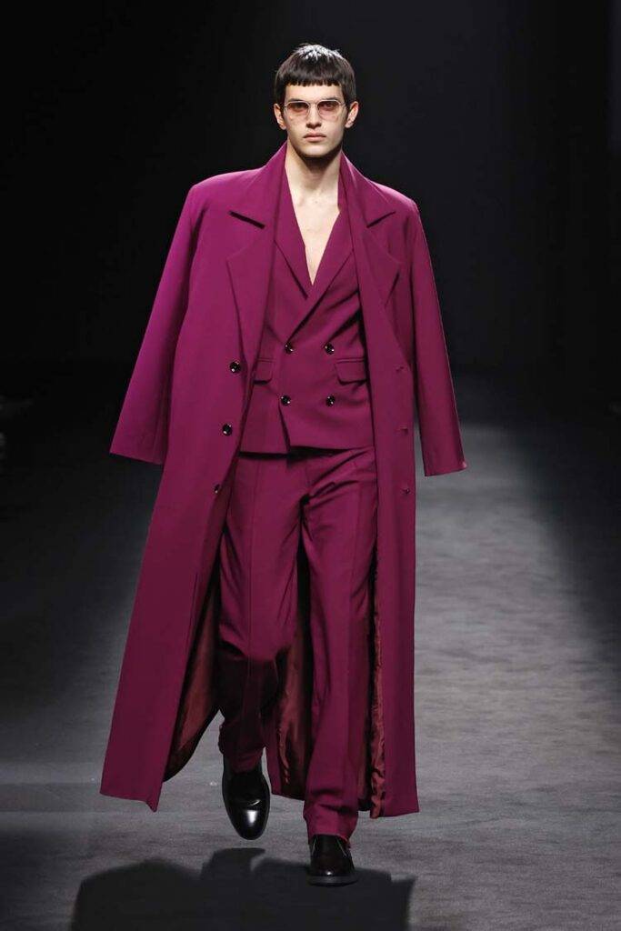 Un modelo masculino viste un traje morado con abrigo largo en una pasarela.