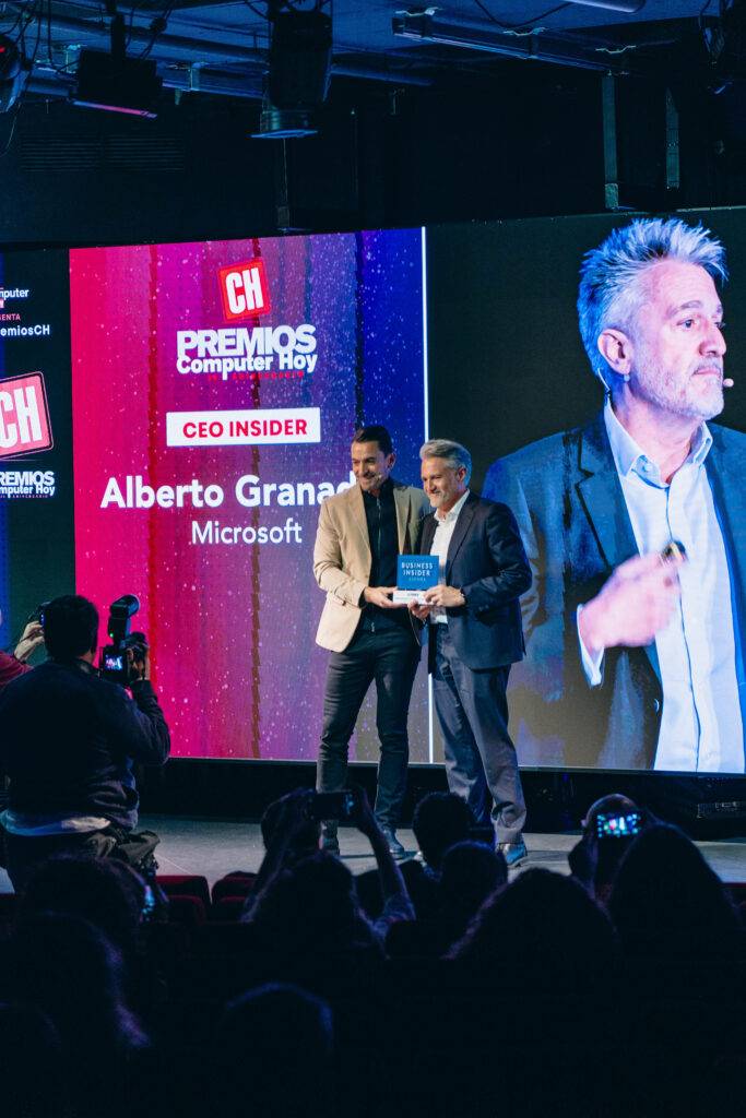 Una ceremonia de premios donde dos personas se dan la mano y uno de ellos sostiene un objeto, con una pantalla en el fondo que muestra información del evento.