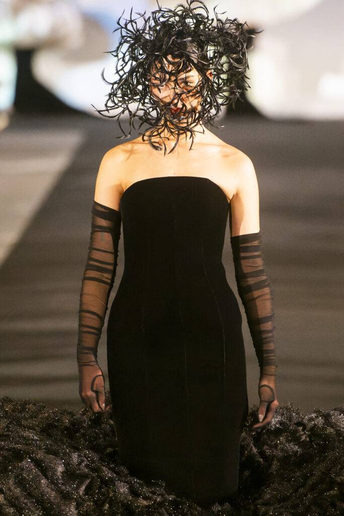 Una modelo presenta un vestido negro ajustado en una pasarela, con un sombrero compuesto de hebras oscuras.