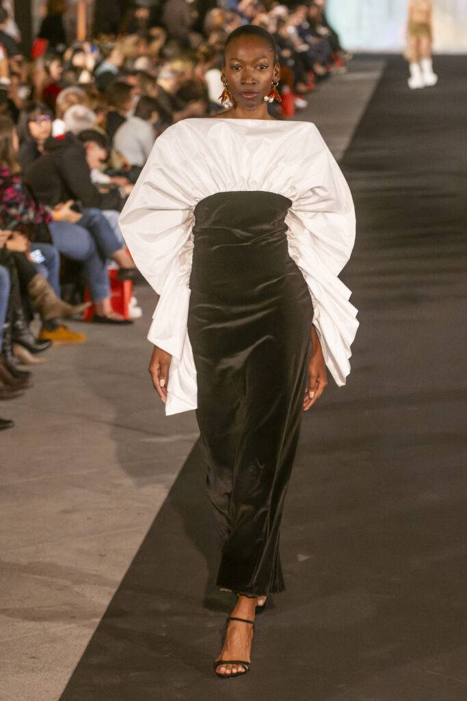 Una modelo camina por la pasarela con un vestido elegante de dos tonos mientras el público observa.