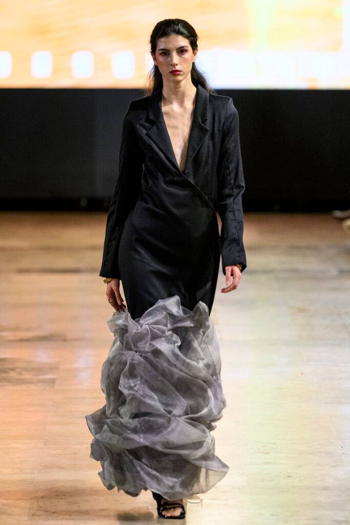 La imagen muestra a una modelo caminando por la pasarela con un vestido largo y elegante que tiene una falda de capas de tejido traslúcido.