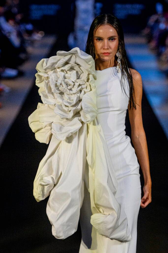 Modelo caminando por la pasarela con un vestido blanco y una gran flor blanca como accesorio.