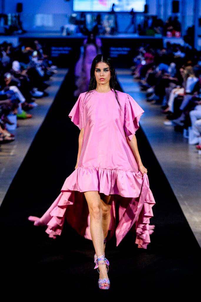 Una modelo camina por la pasarela luciendo un vestido rosa con volantes.
