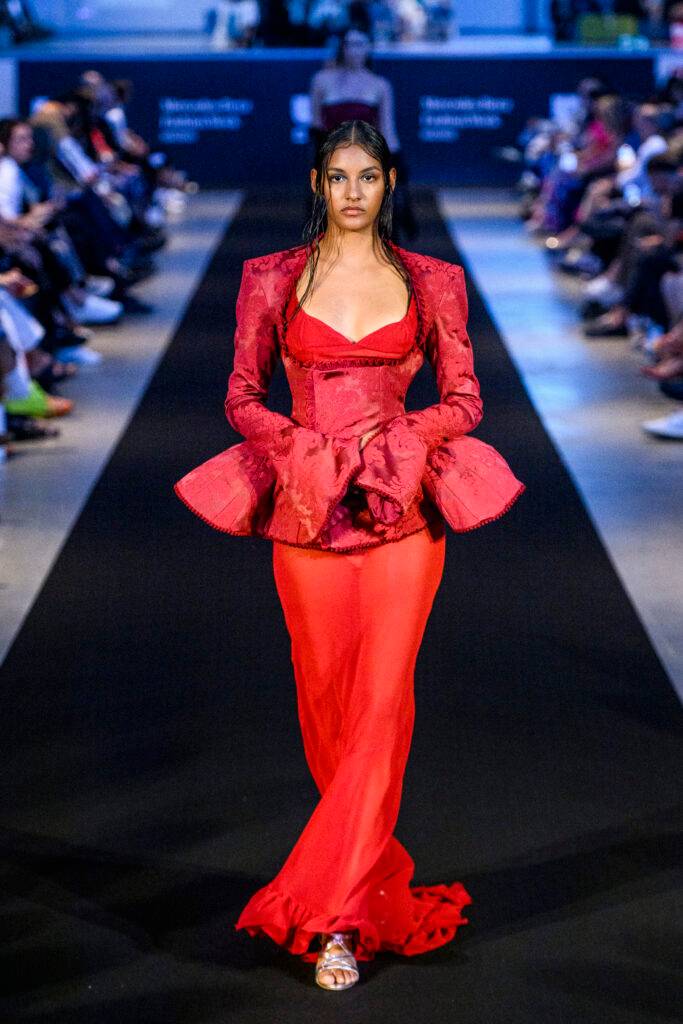Una modelo camina por la pasarela luciendo un vestido rojo elegante con detalles estructurados.