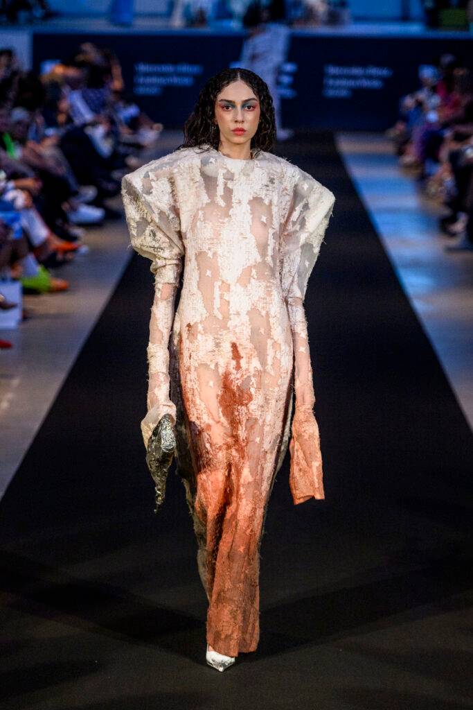 Una modelo desfila en una pasarela luciendo un vestido de diseño innovador con texturas únicas y colores tierra.