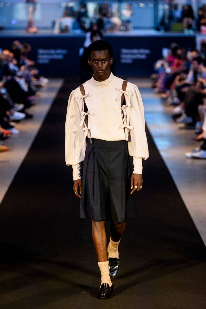 Un modelo camina por la pasarela vistiendo una blusa blanca con detalles y pantalones oscuros en un desfile de moda.