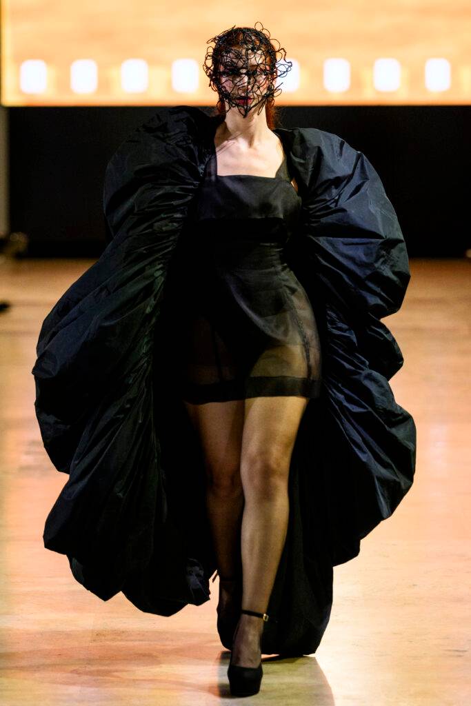 Una modelo camina con un vestido negro de gran volumen y un accesorio en la cabeza que cubre su rostro.
