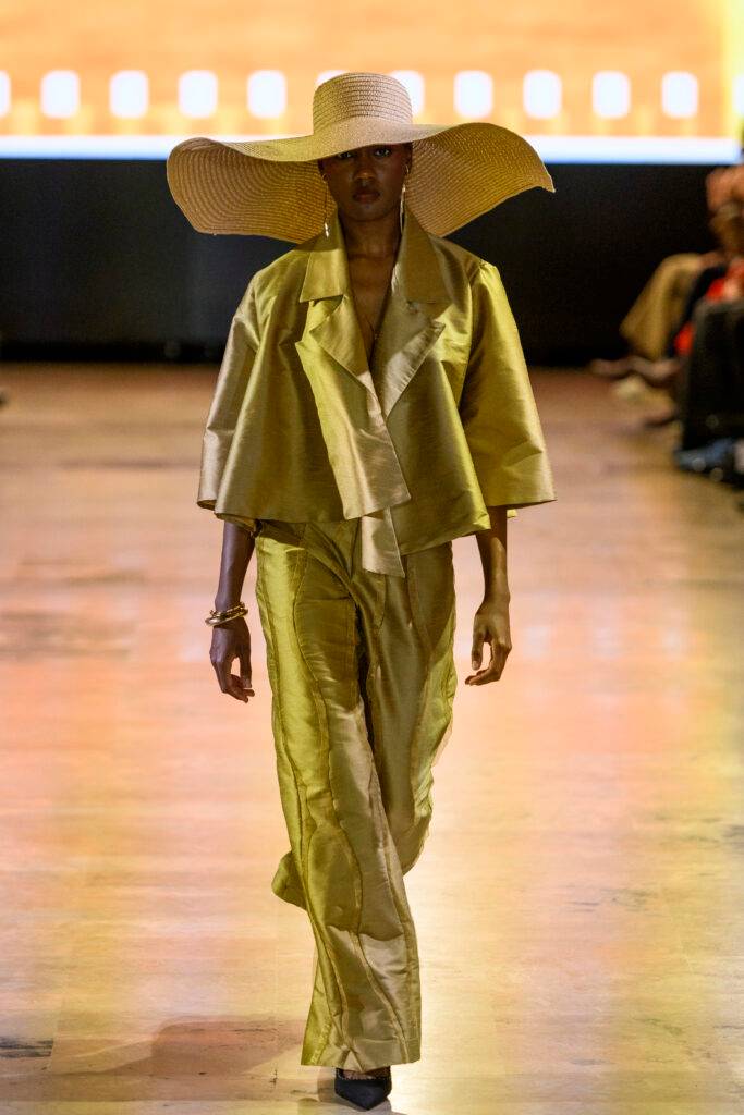 Una modelo camina por la pasarela luciendo un conjunto amarillo y un sombrero amplio.