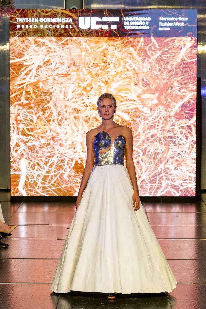 Una modelo presenta un vestido elegante en un evento de moda, frente a una pantalla artística de fondo.