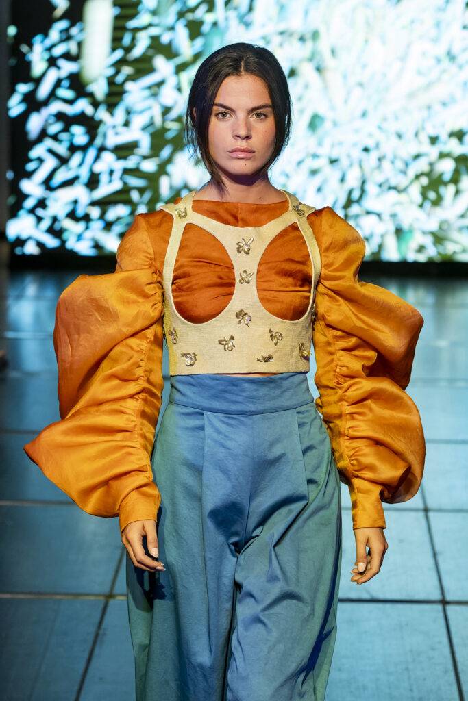 Una modelo presenta un atuendo vanguardista con una blusa de mangas grandes y pantalones holgados en un entorno de pasarela.