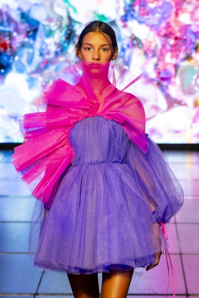 Una modelo presenta un vestido de tul color púrpura y rosa en un entorno artístico y colorido.