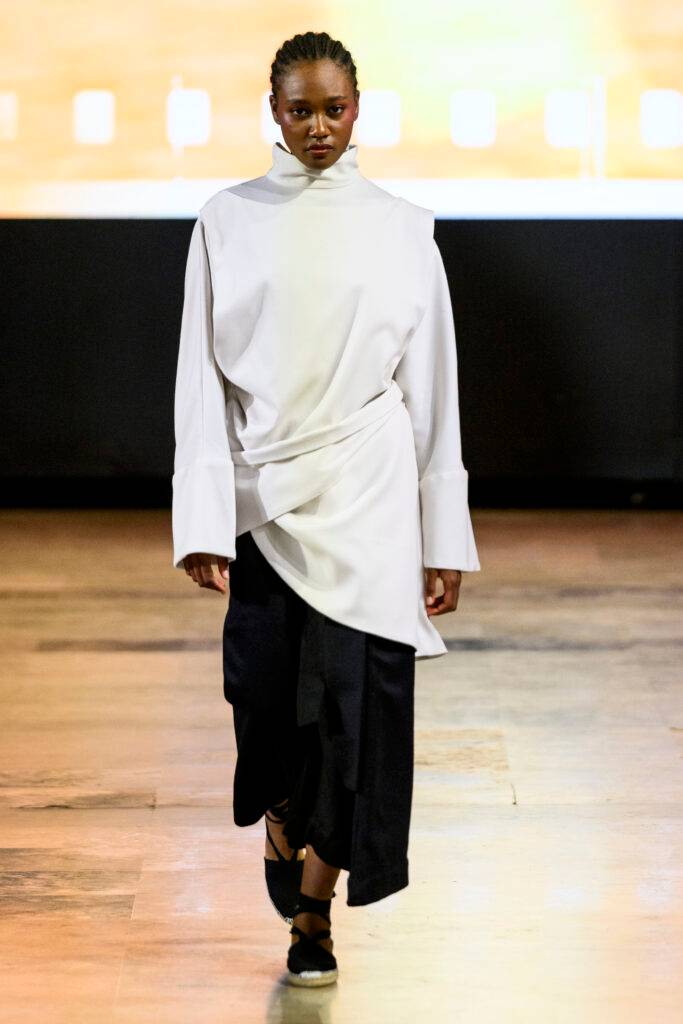 Una modelo camina por la pasarela luciendo una vestimenta elegante en tonos claros y oscuros.