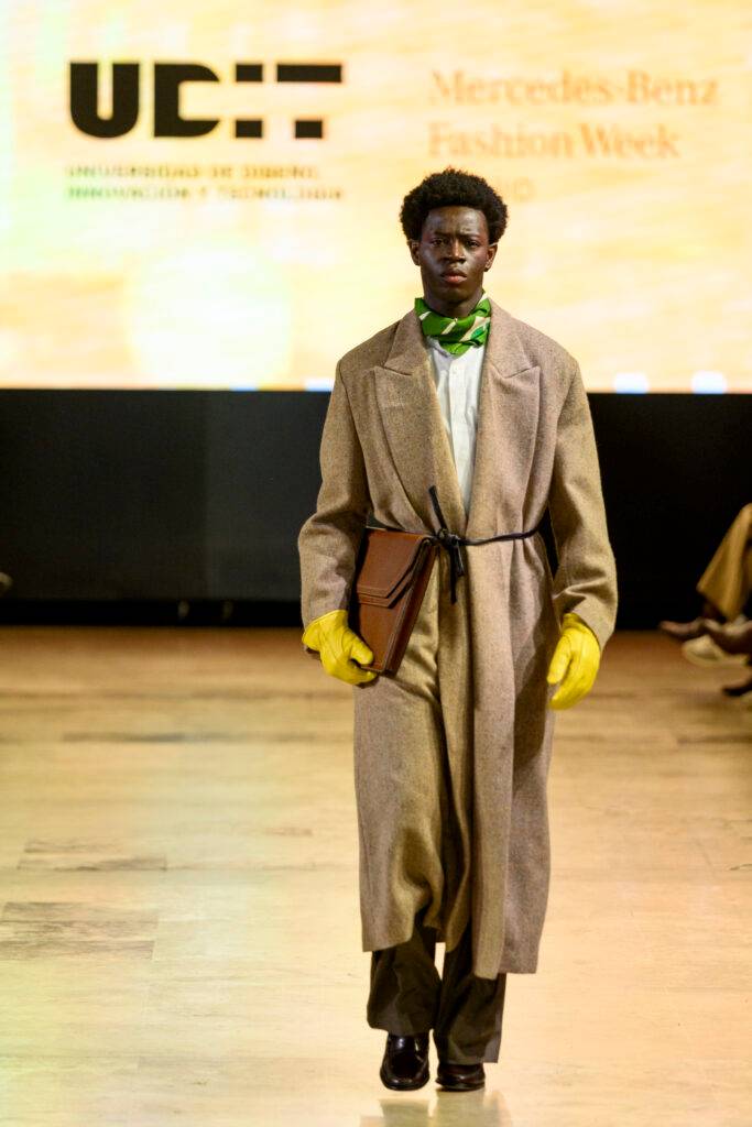 Un modelo caminando por la pasarela durante la Semana de la Moda de Mercedes-Benz, luciendo un abrigo largo y guantes amarillos.