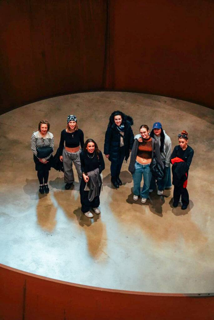 Grupo de siete mujeres posando en un círculo interior con fondo naranja.