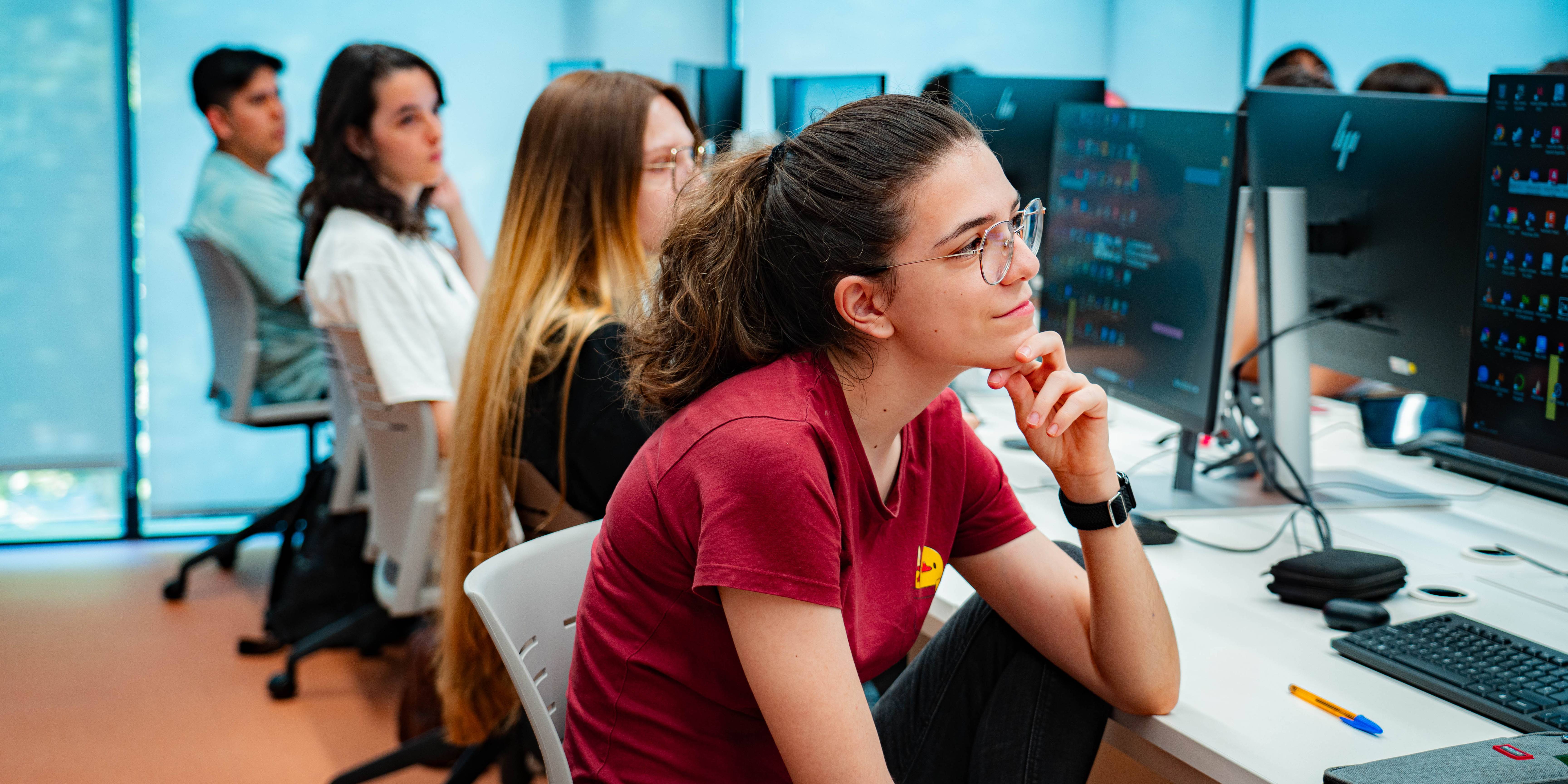 Un grupo de estudiantes se encuentra en una sala de computadoras, prestando atención a una clase.