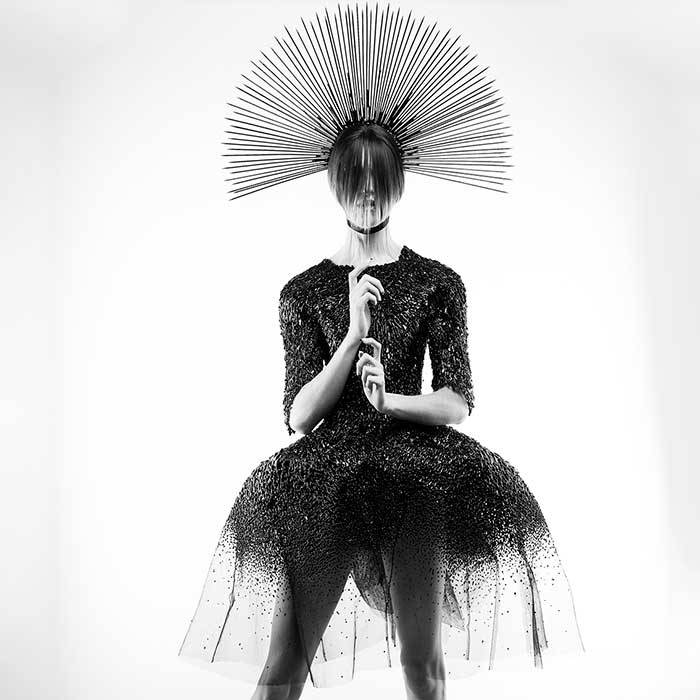 Una modelo lleva un vestido oscuro y voluminoso, con un tocado de varillas en la cabeza y un aspecto enigmático.