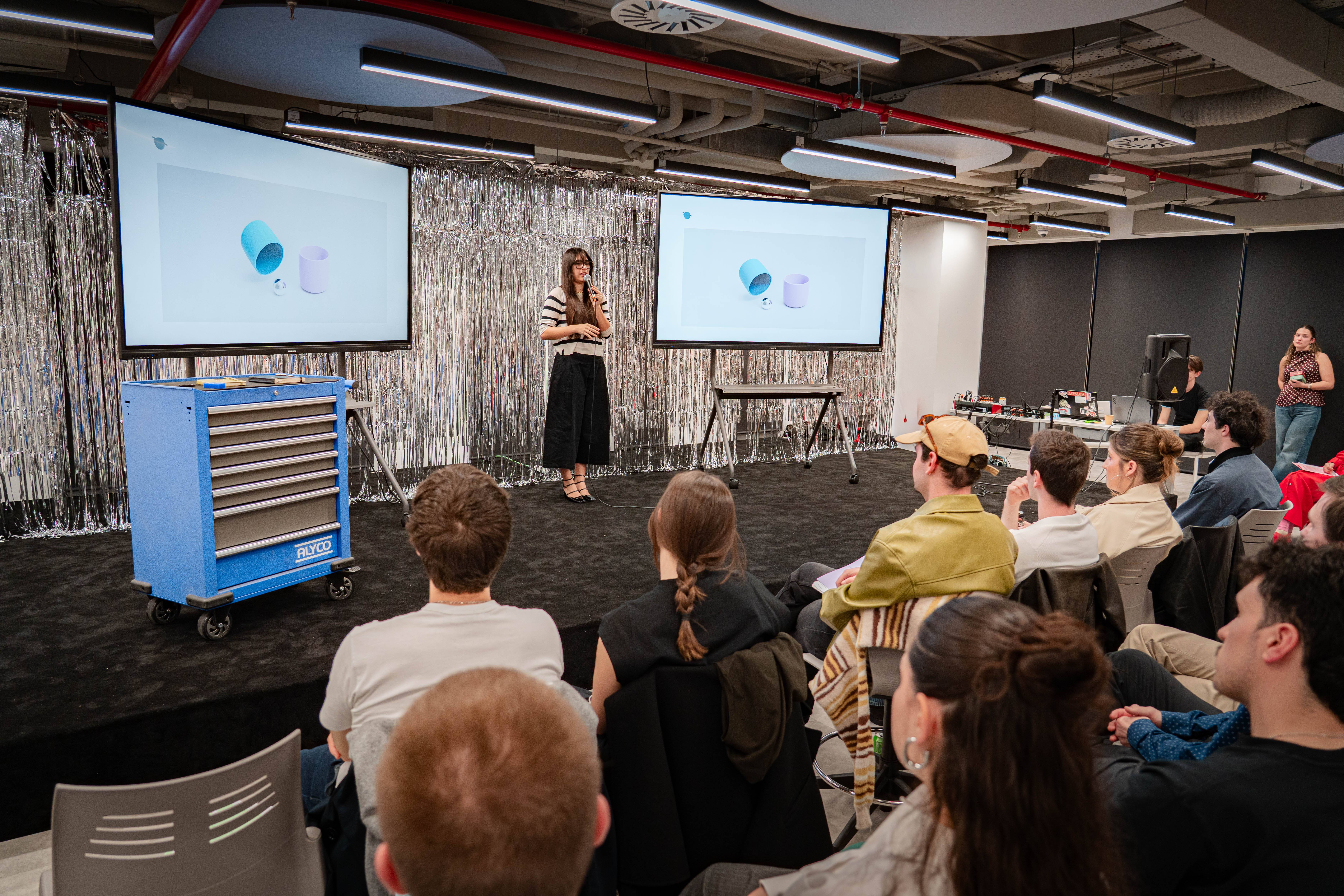 Una presentadora está hablando frente a un público en un evento, con pantallas que muestran una presentación en el fondo.