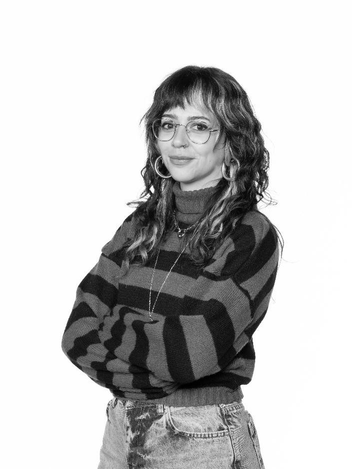 Una persona sonriente con un suéter a rayas y gafas, de pie con los brazos cruzados en un fondo blanco.