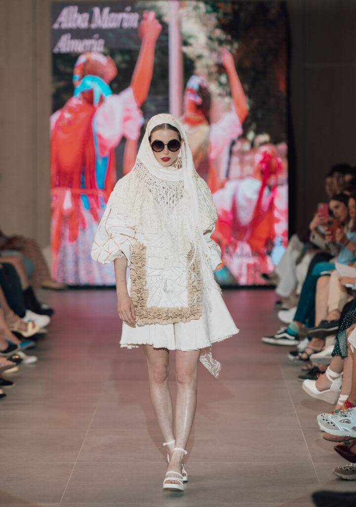 Una modelo camina por la pasarela luciendo un vestido blanco con detalles en crochet y un velo, mientras una multitud observa.
