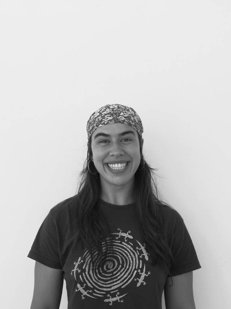Una mujer sonriente con una camiseta oscura y un pañuelo en la cabeza, frente a un fondo blanco.