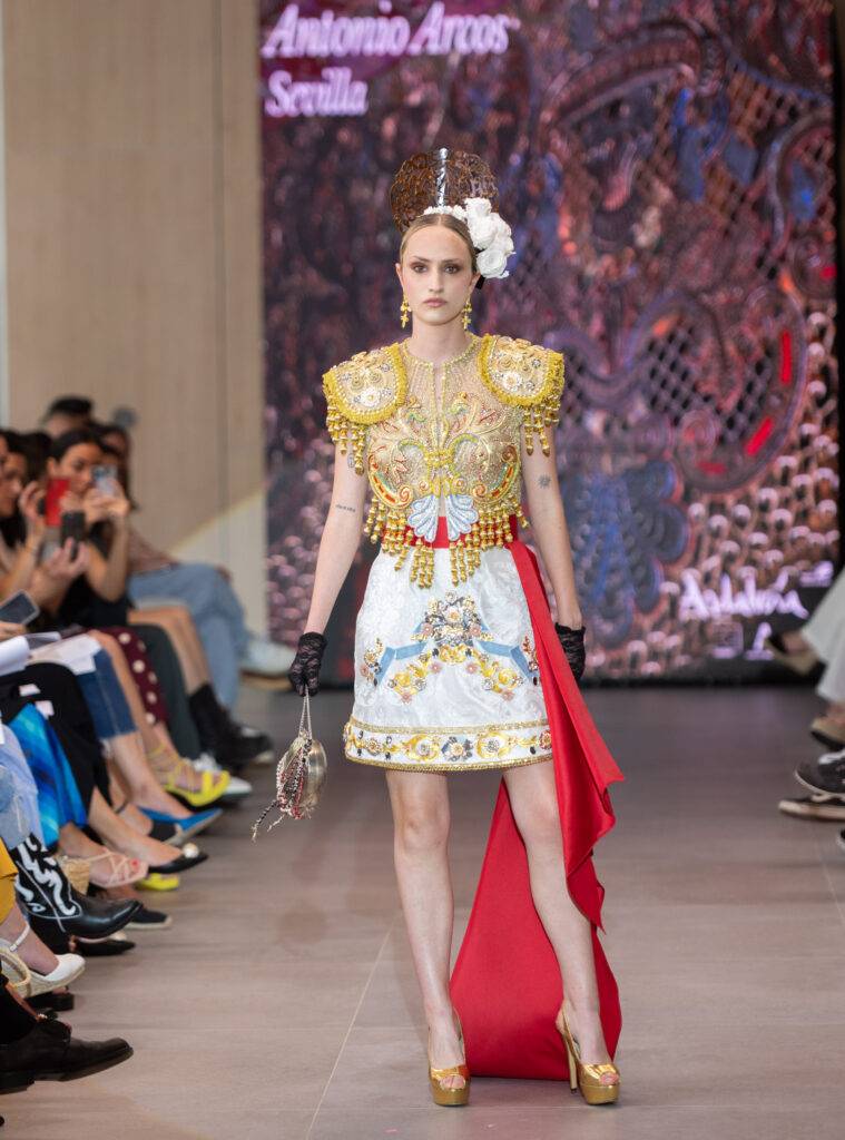 Un modelo desfilando en una pasarela con un vestido inspirado en la cultura andaluza, adornado con elementos tradicionales y contemporáneos.