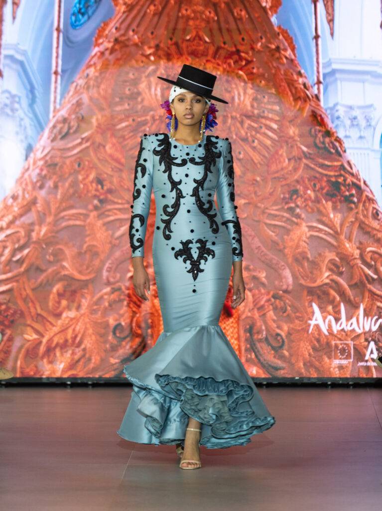 Una modelo camina por la pasarela luciendo un vestido largo de color azul con detalles negros y un sombrero.