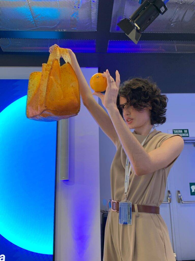 Una joven sostiene una bolsa de color naranja y una naranja en un entorno de oficina.