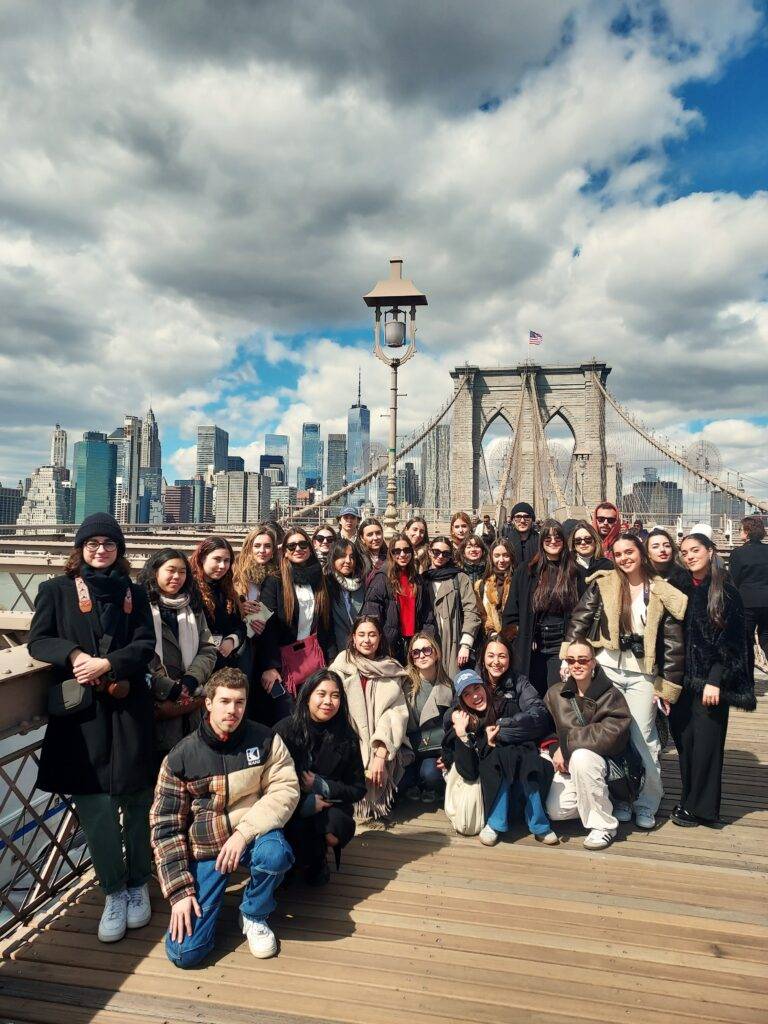Grupo de personas posando en el puente de Brooklyn con un fondo de rascacielos y nubes.