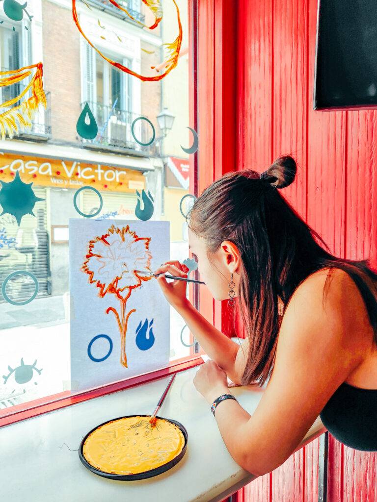 Una mujer está pintando una flor en una hoja frente a una ventana con un fondo colorido.