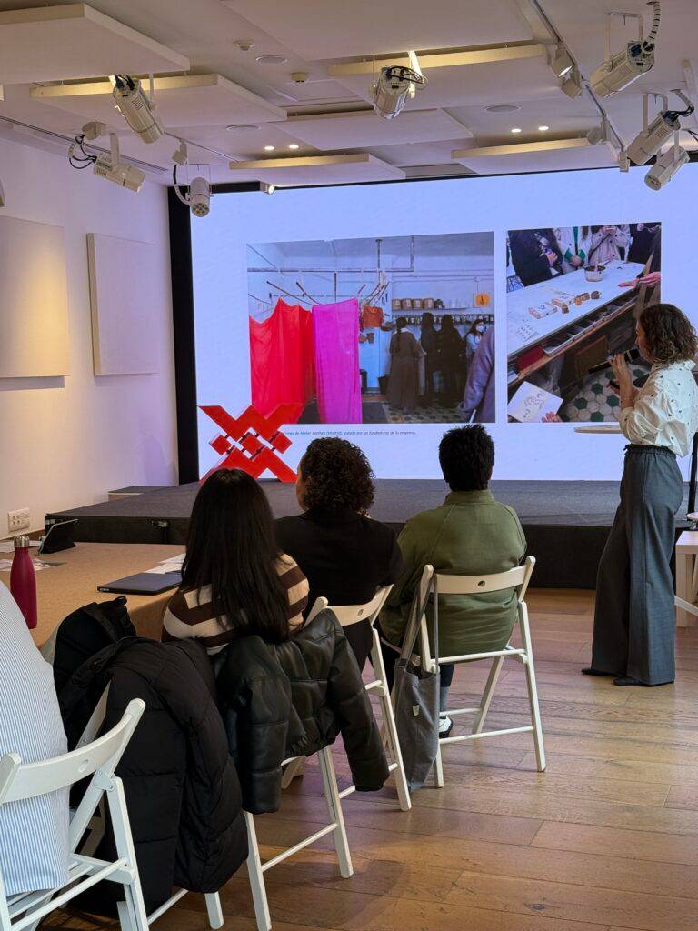 Una presentación en un evento con un proyector mostrando imágenes mientras la presentadora habla ante un grupo de asistentes.