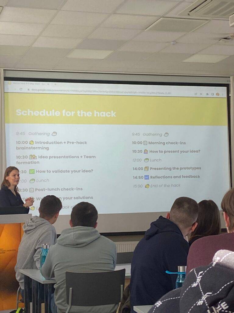 Una presentadora expone el cronograma de un hackathon a un grupo de personas en un aula.