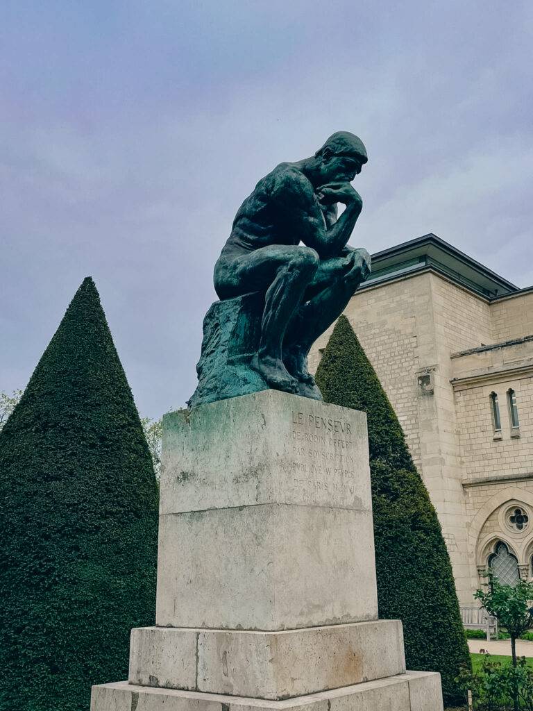 Escultura del pensador en un jardín con árboles podados.