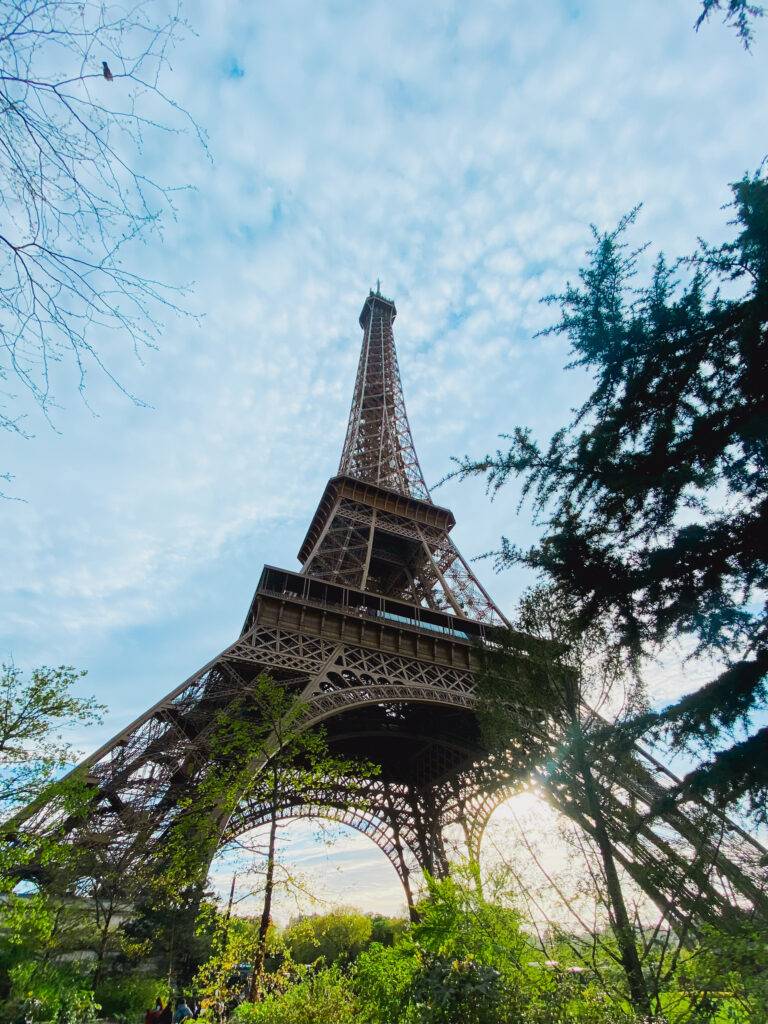 La imagen muestra la Torre Eiffel rodeada de árboles y un cielo parcialmente nublado.