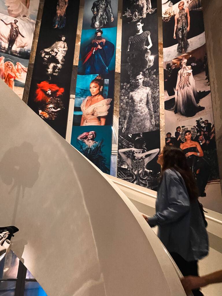 Una persona sube una escalera en un museo que exhibe fotografías de trajes de alta costura.