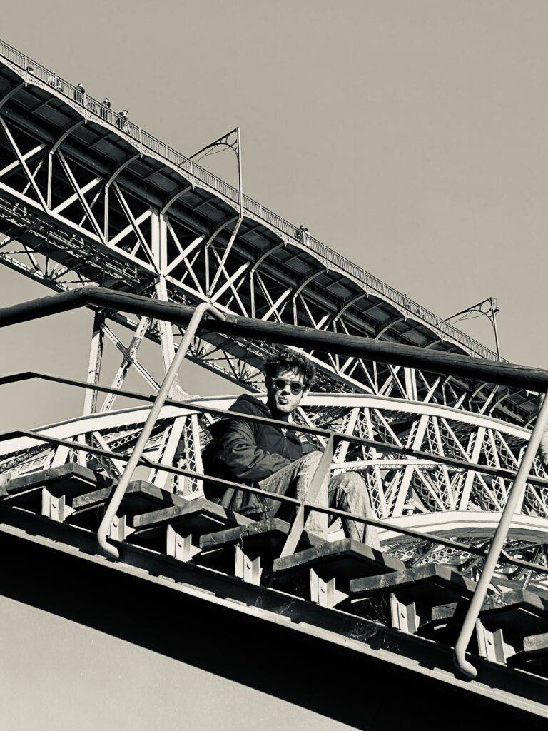 Una persona sonriente sentada en una escalera, con un puente metálico al fondo.