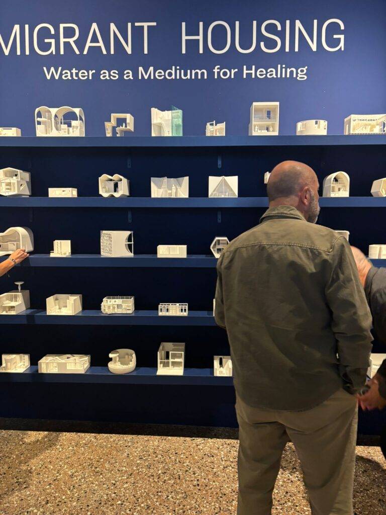 La imagen muestra una exposición de modelos arquitectónicos sobre viviendas para migrantes, con un hombre observando las obras en una pared azul.